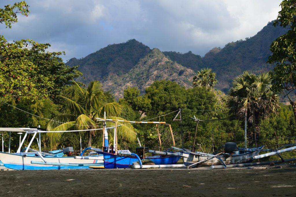 Plage de sable noir de Pemuteran avec bateaux traditionnels en premier plan et montagnes verdoyantes en arrière plan
